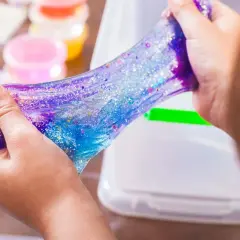 Dziecko bawi się błyszczącym, galaktycznym slime. W tle widać pojemniki z kolorowymi składnikami do slime, przepis na zabawę.