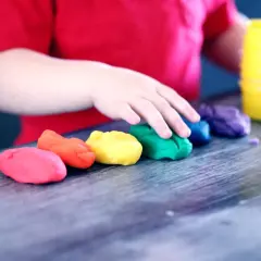 Dziecko w czerwonej koszulce bawi się kolorowymi masami plastycznymi ułożonymi w tęczę.