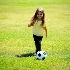 Uśmiechnięta dziewczynka w żółtej bluzce i granatowych spodniach bawi się piłką na trawie. To idealny przykład śmiesznych konkurencji sportowych dla dzieci.