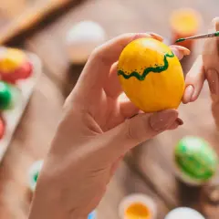 Ręce malują zielone wzory na żółtym jajku, obok leżą inne, barwione tradycyjnie w łupinach cebuli.