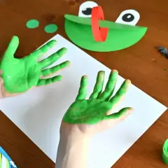 Dziecko maluje zieloną farbą ręce, obok leży papierowa żaba i pędzel.
