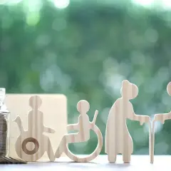 Skarbonka z monetami i drewniane figurki symbolizujące osoby starsze i niepełnosprawne. Czy świadczenie wspierające wlicza się do dochodu?