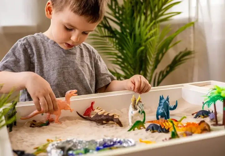 Chłopiec bawi się piaskiem i figurkami dinozaurów. Zabawa sensoryczna cele rozwoju wyobraźni i motoryki małej.