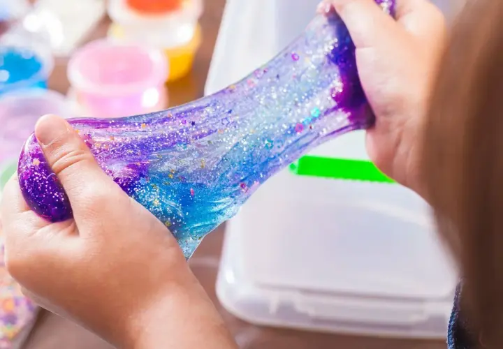 Dziecko bawi się błyszczącym, galaktycznym slime. W tle widać pojemniki z kolorowymi składnikami do slime, przepis na zabawę.