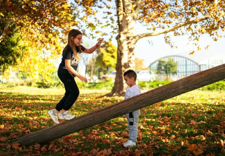 Dziewczynka i chłopiec pokonują drewnianą belkę, jeden z przykładów toru przeszkód dla dzieci. Jesienne liście wokół.