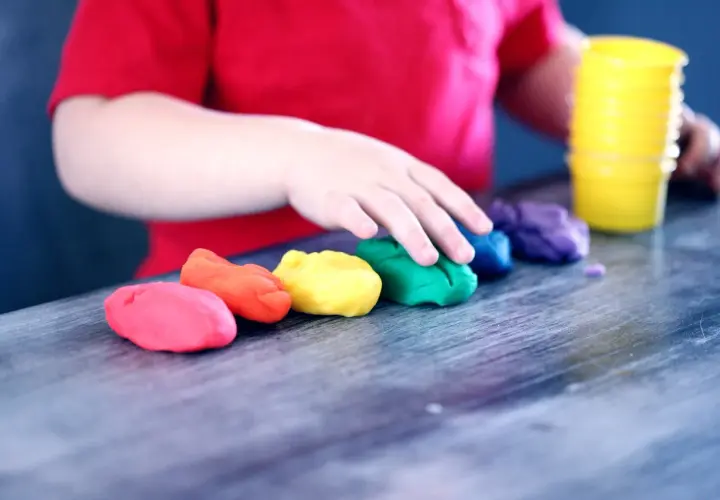 Dziecko w czerwonej koszulce bawi się kolorowymi masami plastycznymi ułożonymi w tęczę.