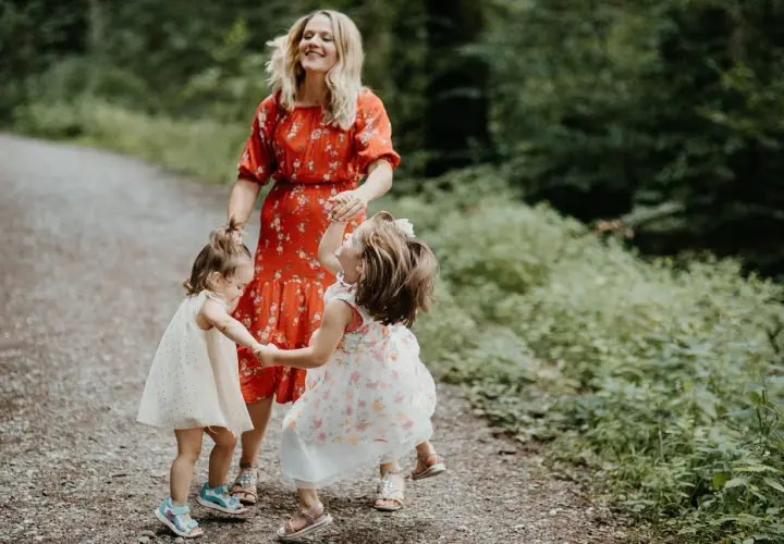 Mama w czerwonej sukience śmieje się, trzymając za ręce dwie córeczki. Dzieci wirują w radosnym tańcu, tworząc ciekawe zabawy taneczne na leśnej ścieżce.