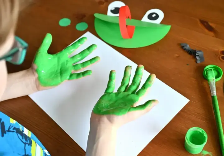 Dziecko maluje zieloną farbą ręce, obok leży papierowa żaba i pędzel.