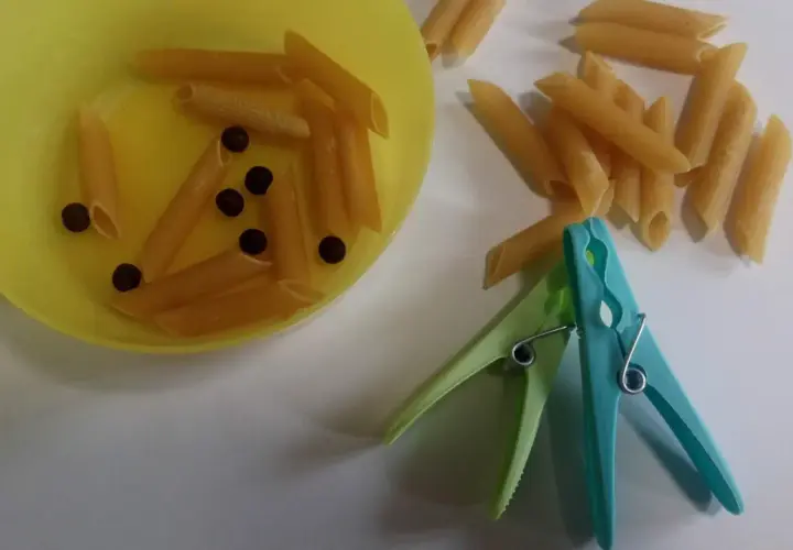 Kolorowe spinacze i makaron penne do ćwiczeń motoryki małej. Dziecko może wkładać makaron do miseczki, używając spinaczy.