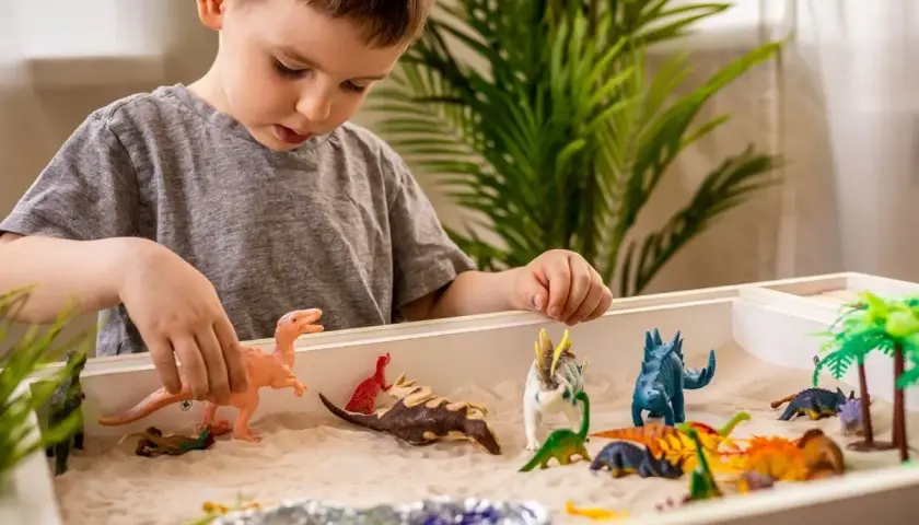 Chłopiec bawi się piaskiem i figurkami dinozaurów. Zabawa sensoryczna cele rozwoju wyobraźni i motoryki małej.