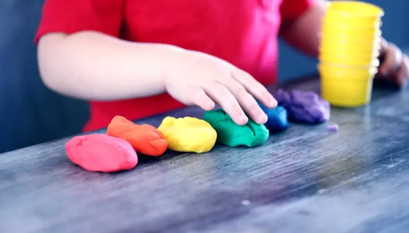 Dziecko w czerwonej koszulce bawi się kolorowymi masami plastycznymi ułożonymi w tęczę.
