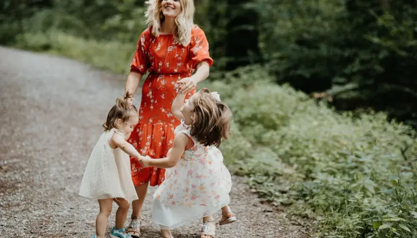 Mama w czerwonej sukience śmieje się, trzymając za ręce dwie córeczki. Dzieci wirują w radosnym tańcu, tworząc ciekawe zabawy taneczne na leśnej ścieżce.