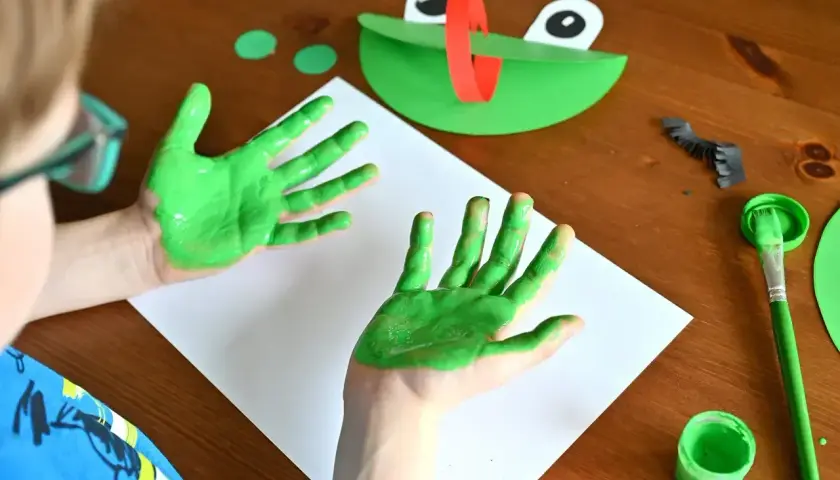 Dziecko maluje zieloną farbą ręce, obok leży papierowa żaba i pędzel.