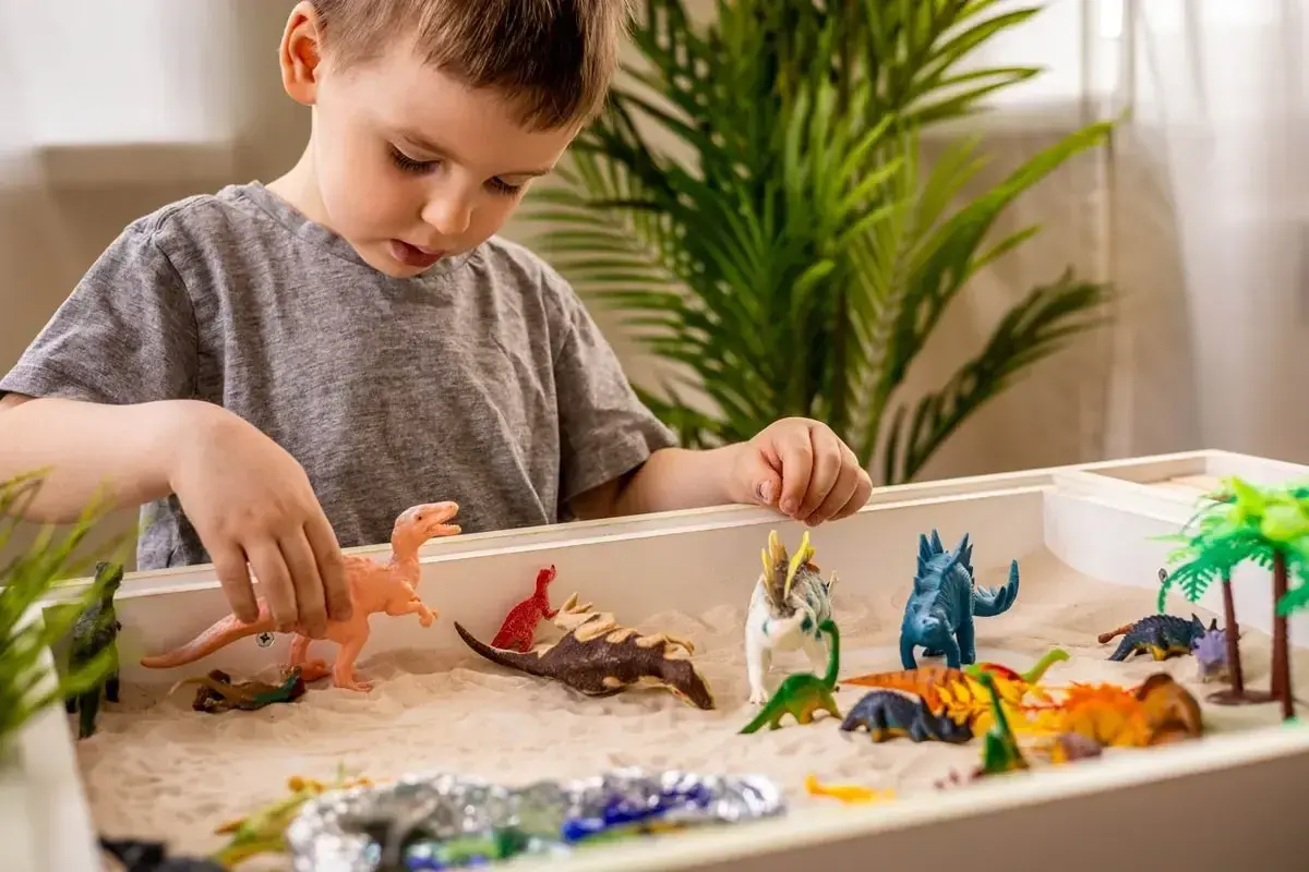 Chłopiec bawi się piaskiem i figurkami dinozaurów. Zabawa sensoryczna cele rozwoju wyobraźni i motoryki małej.