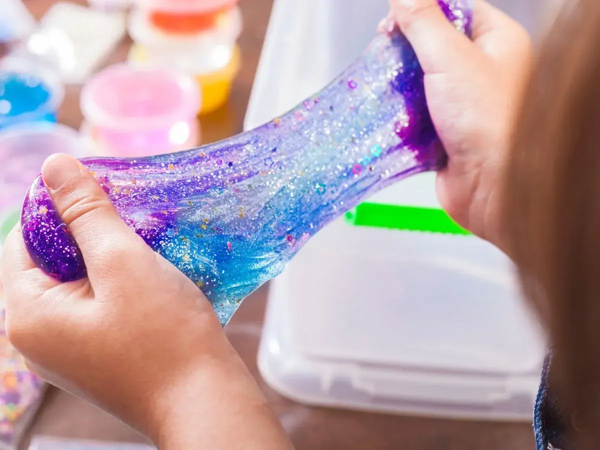 Dziecko bawi się błyszczącym, galaktycznym slime. W tle widać pojemniki z kolorowymi składnikami do slime, przepis na zabawę.