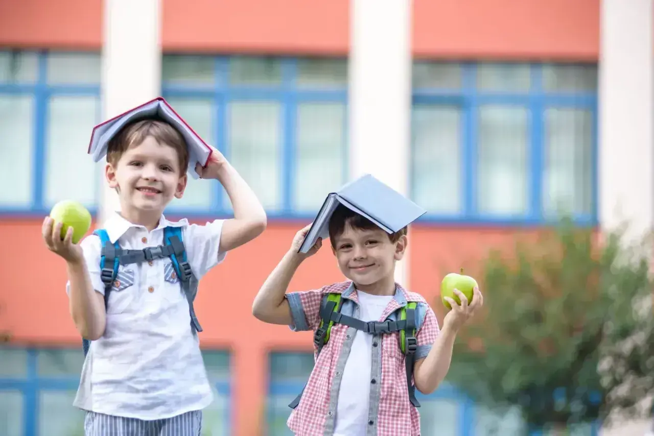 Dwóch chłopców z plecakami i jabłkami w rękach, z książkami na głowach, radośnie wita nowy dzień, kiedy dzieci idą do szkoły.