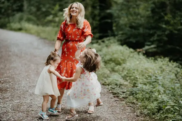 Mama w czerwonej sukience śmieje się, trzymając za ręce dwie córeczki. Dzieci wirują w radosnym tańcu, tworząc ciekawe zabawy taneczne na leśnej ścieżce.